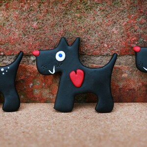 May include: Three black clay sculptures of dogs with white eyes and red accents. The dogs are wearing different accessories, including a bow tie, a heart, and a collar.