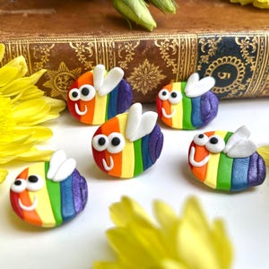 Puede incluir: Un conjunto de decoraciones hechas a mano con forma de abeja. Cada abeja presenta un cuerpo a rayas de arcoíris, alas blancas y una cara sonriente. Las abejas están dispuestas sobre una superficie blanca, rodeadas de flores amarillas.