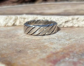 May include: A silver-colored ring with a textured, braided design. The ring is circular and appears to be made of metal. The ring is displayed on a light brown surface with a neutral background.