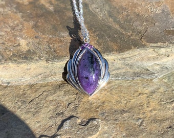May include: A silver necklace with a purple gemstone pendant. The pendant is set in a silver frame with a scalloped edge. The gemstone is surrounded by smaller purple gemstones.
