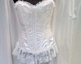 May include: White satin corset with lace detailing and a ruffled hem. The corset is strapless and has a boned bodice.