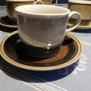 Puede incluir: Un juego de cuatro tazas de café de cerámica marrón y blanco con platillos a juego. Las tazas tienen un acabado esmaltado y una forma redondeada. Los platillos tienen un borde marrón y un centro blanco.
