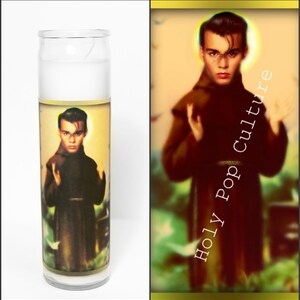 May include: A white candle with a printed image of Johnny Depp dressed as a monk. The image is surrounded by a gold border and the text "Holy Pop Culture" is printed vertically on the right side of the candle.