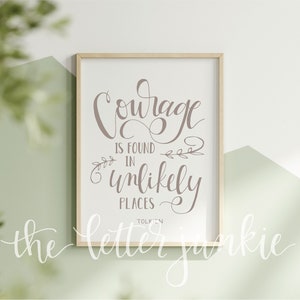May include: A framed print with a quote in cursive script: "Courage is found in unlikely places - Tolkien". The print is on a white background and has a light brown frame. The print is hanging on a wall with a light green background.