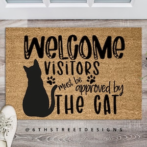 May include: Welcome visitors must be approved by the cat doormat with a black silhouette of a cat. The doormat is made of coir and has a brown background. The text is in black and white.