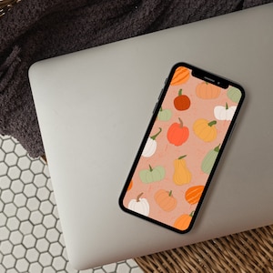 Op de afbeelding: Een laptop met een telefoon die een digitale illustratie van een patroon van oranje, witte en groene pompoenen op een perzikkleurige achtergrond weergeeft.