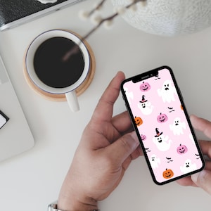 May include: A smartphone with a pink background and a Halloween-themed wallpaper featuring ghosts, pumpkins, and bats.