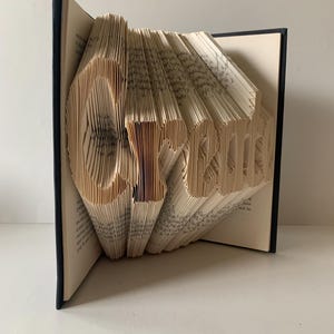 May include: A hardback book with black covers, featuring pages folded to form the word Create. The cream-colored paper pages are precisely manipulated to create a three-dimensional typographic sculpture that stands upright on a flat surface.