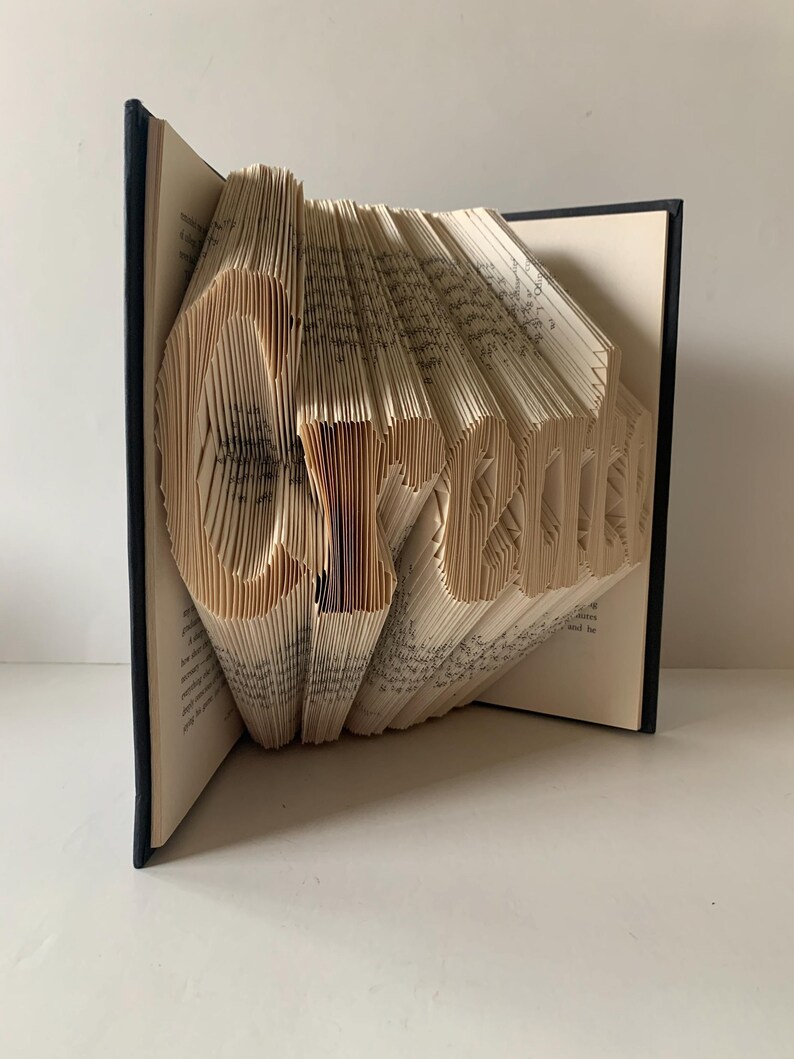 May include: A hardback book with black covers, featuring pages folded to form the word Create. The cream-colored paper pages are precisely manipulated to create a three-dimensional typographic sculpture that stands upright on a flat surface.