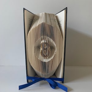 Puede incluir: Una escultura de arte de libro de un ojo azul con un iris y una pupila blancos. El libro está doblado en una forma tridimensional y está atado con una cinta azul.