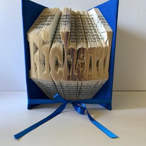 May include: A folded book art sculpture with the name Richard formed by the precisely creased pages. The book is held open by bright blue covers and features a matching blue satin ribbon tied in a bow at the base of the pages.