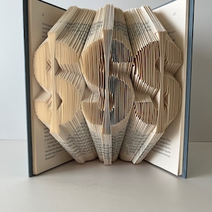 May include: A book with pages folded into the shape of the number 353. The book is open and the pages are folded to create a three-dimensional effect. The book is on a white surface.