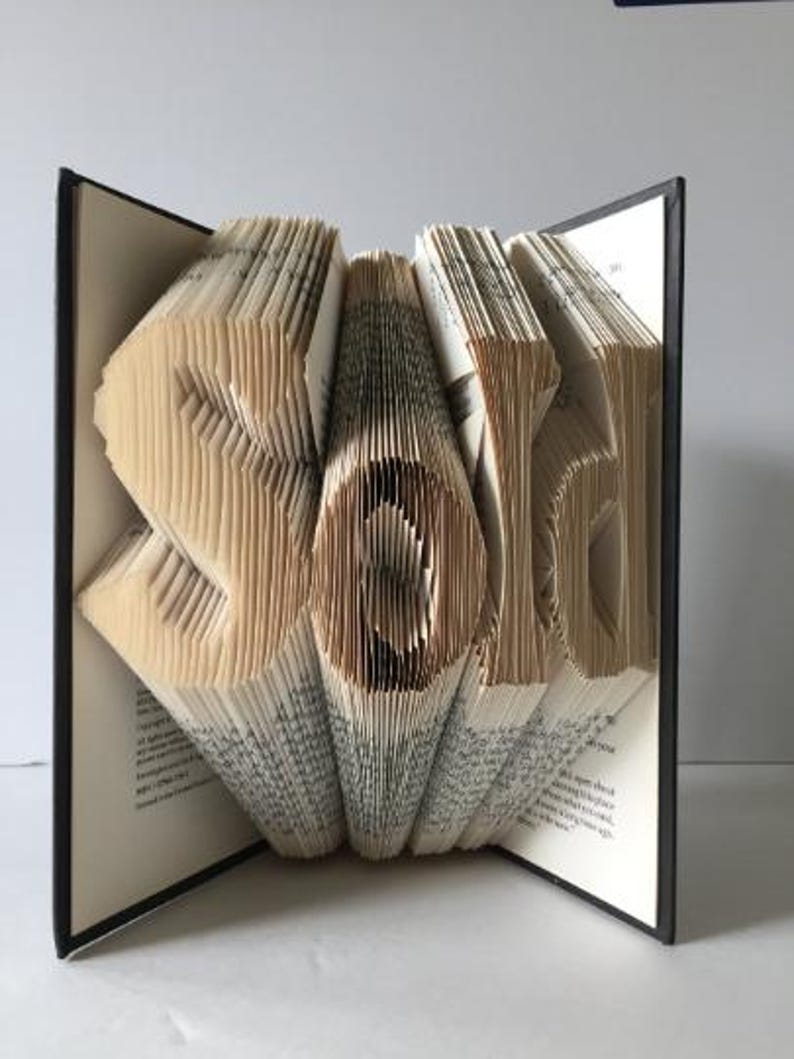 May include: A book art sculpture of the word "Sold" created by folding the pages of a book. The sculpture is displayed open on a black surface.