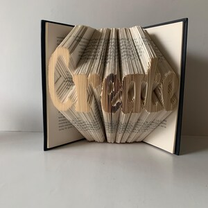 May include: A folded book art sculpture featuring the word Create formed by precisely folding the individual pages of a hardcover book. The black book covers are visible on the sides, while the cream-colored pages are folded to display the text.