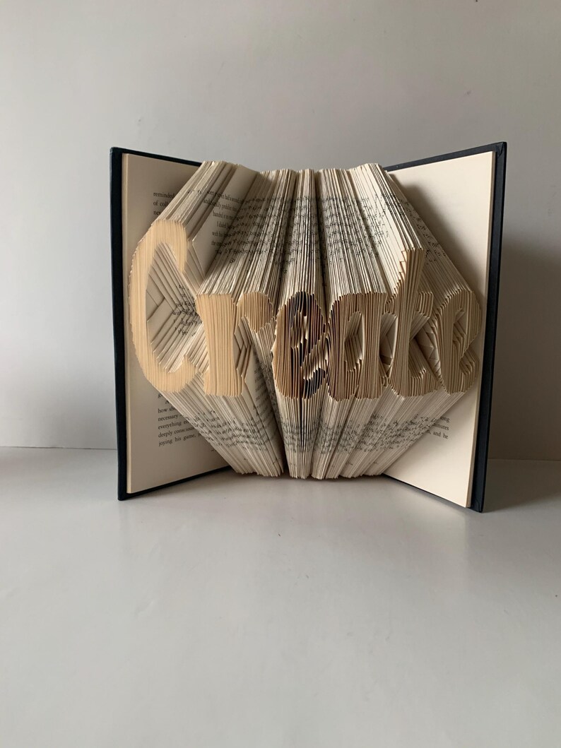 May include: A folded book art sculpture featuring the word Create formed by precisely folding the individual pages of a hardcover book. The black book covers are visible on the sides, while the cream-colored pages are folded to display the text.