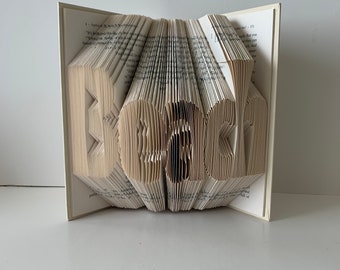 May include: A book art sculpture of the word "Beach" created by folding the pages of a book. The sculpture is made from a paperback book with a white cover.