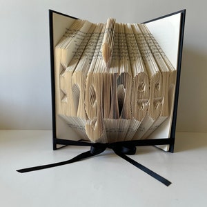 May include: A book art sculpture of the word "Forever" created by folding the pages of a book. The book is bound in black and has a black ribbon tied around it.