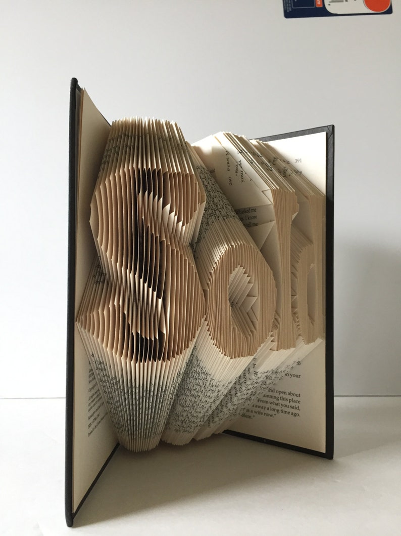 May include: A book art sculpture of the word "Roll" created by folding the pages of a book. The book is bound in black and the pages are a cream color.