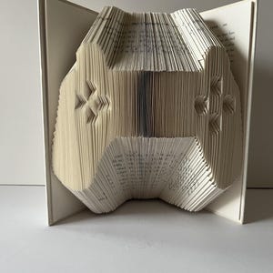 May include: A unique book art piece featuring a folded paper design of a video game controller. The controller shape is created by meticulously folding the pages of an open book, showcasing the intricate details of the design.