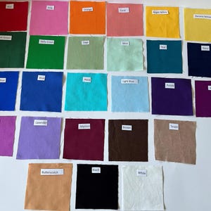May include: A collection of twenty-three square fabric swatches arranged in rows, each featuring a small white label with text identifying the color. The colors include red, pink, orange, coral, bright yellow, banana yellow, dark green, kelly green, sage, mint, teal, navy, royal blue, blue, aqua, light blue, purple, violet, magenta, lavender, maroon, brown, taupe, butterscotch, black, and white.