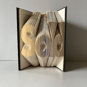 May include: A book art sculpture of the word "sale" created by folding the pages of a book. The book is bound in black and the pages are a cream color.
