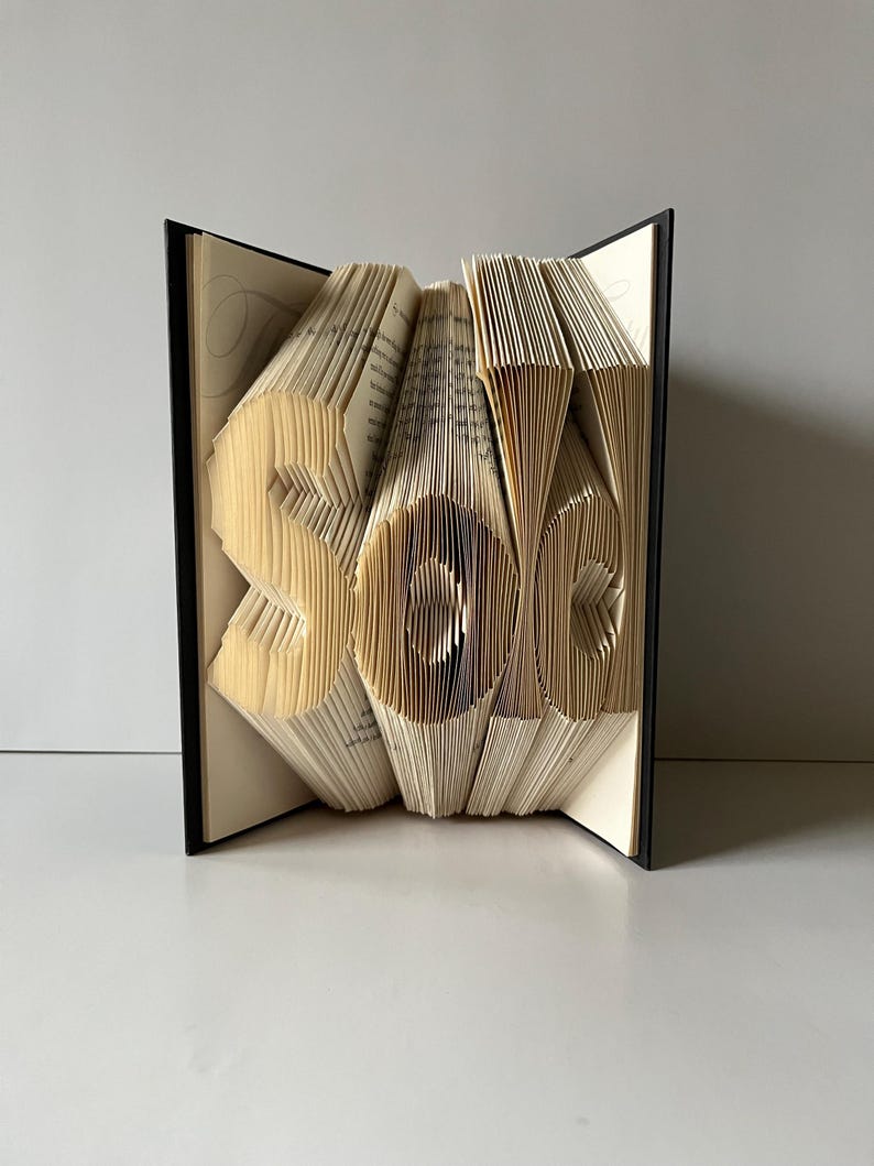 May include: A book art sculpture of the word "sale" created by folding the pages of a book. The book is bound in black and the pages are a cream color.