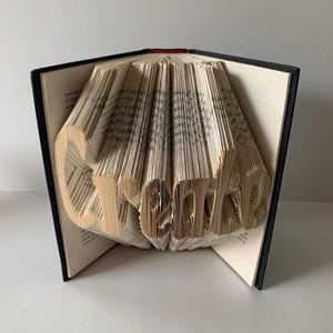 May include: A hardback book with black covers, featuring pages folded to form the word Create. The cream-colored paper edges are precisely manipulated to create three-dimensional lettering that stands out against the dark book spine and covers.