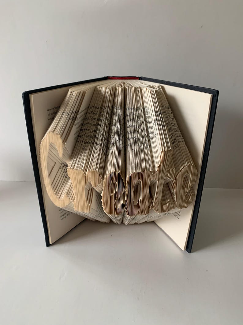 May include: A hardback book with black covers, featuring pages folded to form the word Create. The cream-colored paper edges are precisely manipulated to create three-dimensional lettering that stands out against the dark book spine and covers.