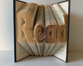 May include: A book with the word "Read" folded into the pages. The book is open and the pages are folded to create the letters. The book is black with a white spine.