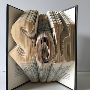 May include: A book art sculpture of the word "Sold" created by folding the pages of a book. The sculpture is displayed open on a black surface.