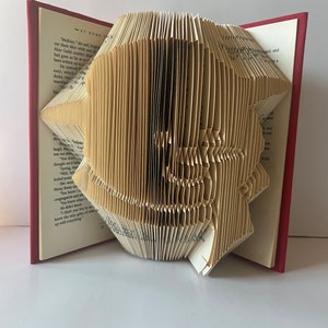 May include: A book with pages folded into a three-dimensional shape, creating a sculpture of a bird. The book is open and the pages are folded in a way that creates a realistic-looking bird. The book is bound in red and the pages are cream-colored.