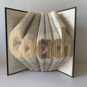 Könnte beinhalten: Eine Buchkunstskulptur des Wortes "Coach", die durch das Falten der Seiten eines Buches entstanden ist. Das Buch ist geöffnet und die Seiten sind gefaltet, um die Buchstaben zu bilden. Das Buch steht auf einer weißen Oberfläche.