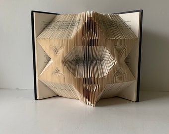 May include: A book art sculpture in the shape of a star, folded from the pages of a book. The book is open and the pages are folded into a three-dimensional star shape.