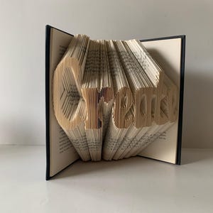 May include: A book sculpture featuring the word Create formed by precisely folded pages. The book has a dark cover and cream-colored paper pages that are manipulated to display the three-dimensional lettering in a clear, legible font style.