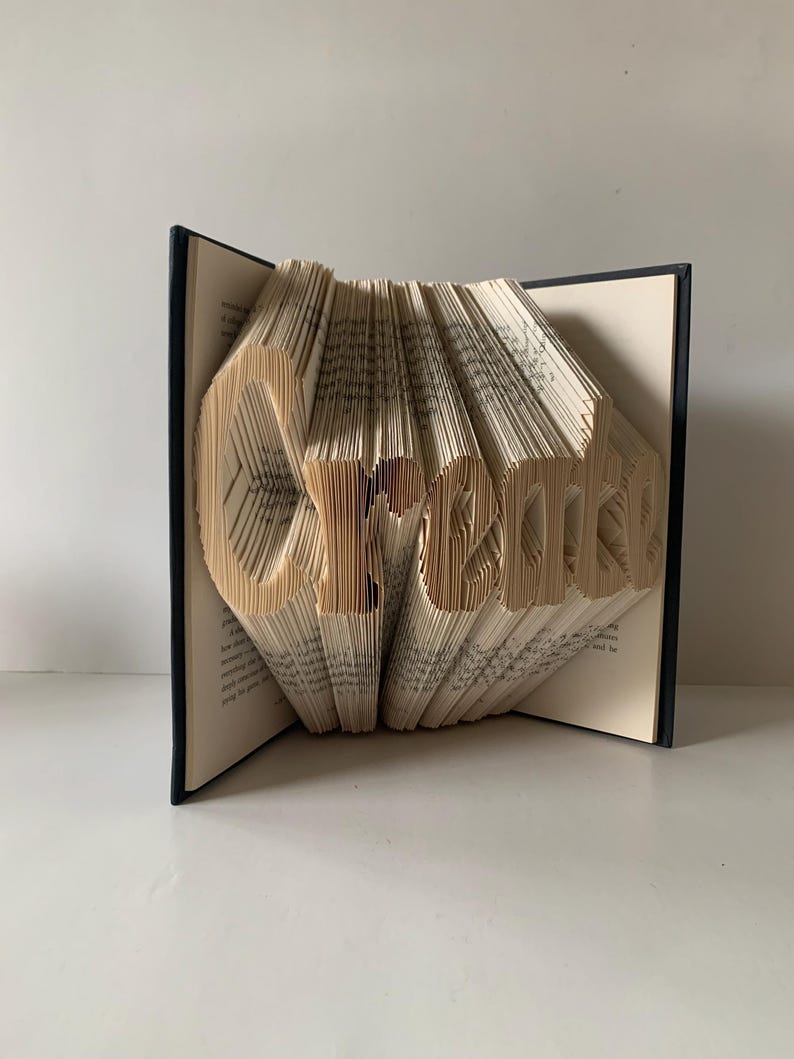 May include: A book sculpture featuring the word Create formed by precisely folded pages. The book has a dark cover and cream-colored paper pages that are manipulated to display the three-dimensional lettering in a clear, legible font style.
