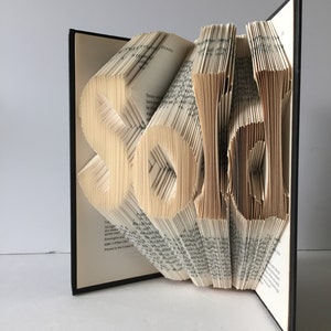 May include: A book art sculpture of the word "sold" created by folding the pages of a book. The book is open and the pages are folded into the shape of the letters. The book is standing on a white surface.