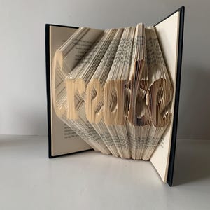 May include: A book art sculpture featuring the word Create formed by precisely folded pages. The book has a black cover and cream-colored paper pages with black printed text. The pages are fanned out to display the three-dimensional lettering.