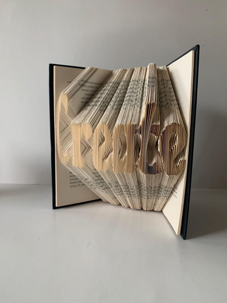 May include: A book art sculpture featuring the word Create formed by precisely folded pages. The book has a black cover and cream-colored paper pages with black printed text. The pages are fanned out to display the three-dimensional lettering.