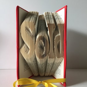May include: A red book with a yellow ribbon is folded into the word "Sold".
