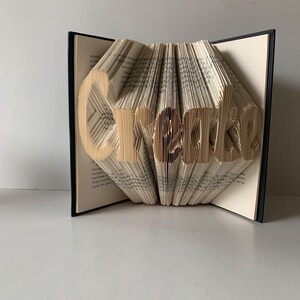 May include: A folded book art sculpture featuring the word Create formed by precisely creased and folded pages. The book has a dark black cover and cream-colored paper pages with visible printed text, standing open to display the three-dimensional lettering.