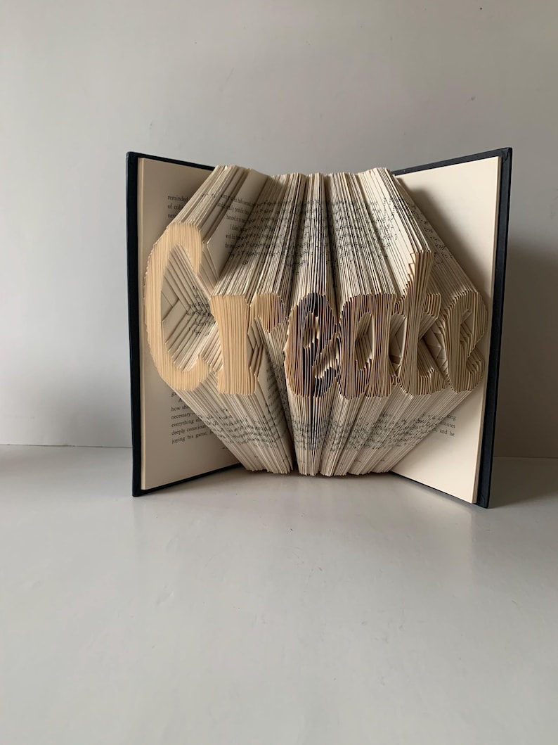 May include: A folded book art sculpture featuring the word Create formed by precisely creased and folded pages. The book has a dark black cover and cream-colored paper pages with visible printed text, standing open to display the three-dimensional lettering.