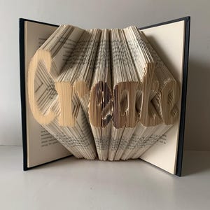 May include: A book with pages folded to form the word Create. The book has a dark cover and cream-colored pages with black printed text. The folded pages create a three-dimensional effect, standing upright with the word clearly visible in the center.