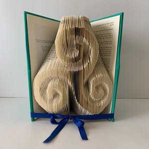 May include: A unique book art sculpture with teal covers and pages folded into a three-dimensional design. A blue ribbon is tied around the book. The book is open, revealing text on the pages.