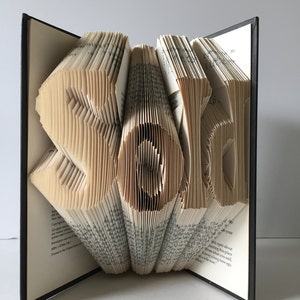 May include: A book art sculpture of the word "Sold" created by folding the pages of a book. The sculpture is made from a vintage book with a black cover.