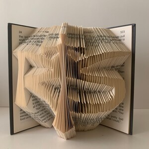Può includere: Un libro piegato in una forma tridimensionale, creando un pezzo d'arte unico e decorativo. Il libro è aperto e le pagine sono piegate in un motivo complesso, formando una forma che ricorda un uccello. Il libro è fatto di carta e ha una copertina marrone.