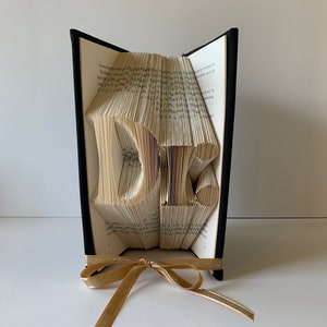 May include: A book art sculpture of the letters "DE" folded from the pages of a book. The book is bound in black fabric and has a gold ribbon tied around it.