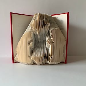 Peut inclure: Un livre rouge dont les pages sont pliées en une sculpture 3D de deux personnages. Le livre est ouvert et la sculpture est au centre de l'image.