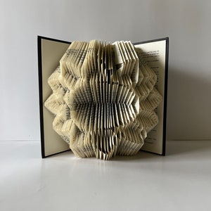 Puede incluir: Una escultura de arte de libro con páginas dobladas en un diseño 3D. El libro está abierto y las páginas están dobladas en un patrón geométrico. El libro está sobre una superficie blanca.