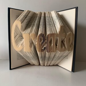 May include: A book sculpture with pages folded to form the word Create. The book has a dark cover and cream-colored pages with visible black text. The pages are fanned out to create a three-dimensional effect, displaying the letters clearly.