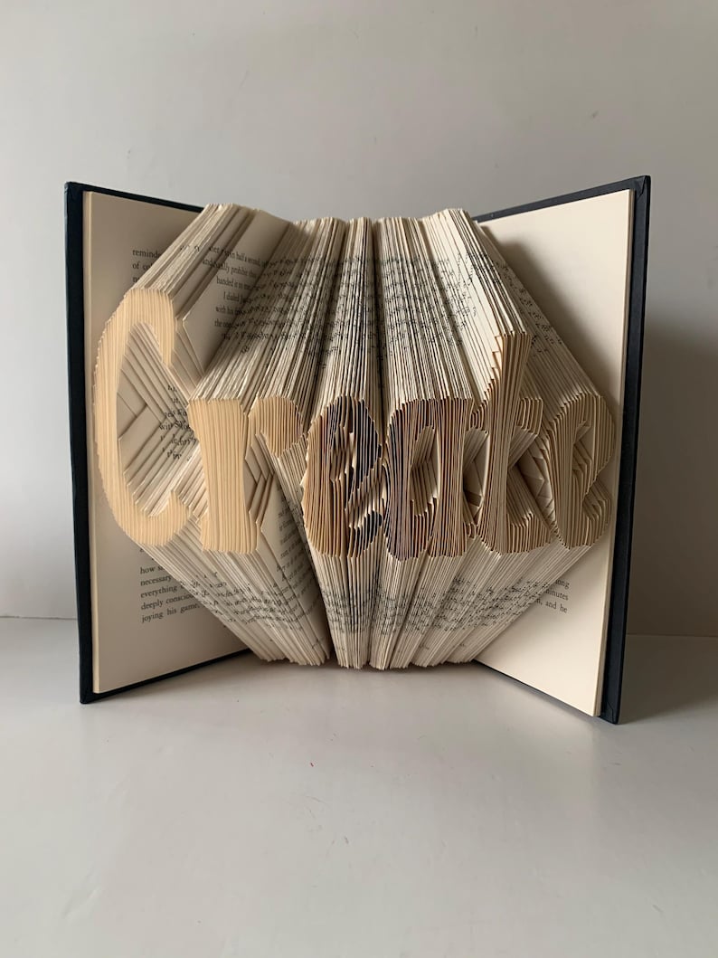 May include: A book sculpture with pages folded to form the word Create. The book has a dark cover and cream-colored pages with visible black text. The pages are fanned out to create a three-dimensional effect, displaying the letters clearly.